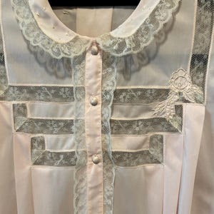 May include: A light pink vintage blouse with a Peter Pan collar and button-up front. The blouse features delicate lace trim on the collar, front, and sleeves, with a floral applique on the chest. The fabric appears to be a soft, lightweight material.