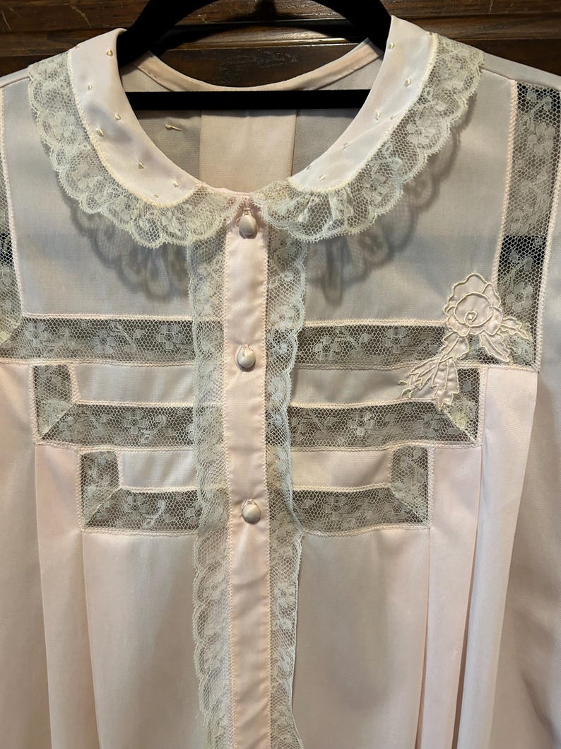 May include: A light pink vintage blouse with a Peter Pan collar and button-up front. The blouse features delicate lace trim on the collar, front, and sleeves, with a floral applique on the chest. The fabric appears to be a soft, lightweight material.