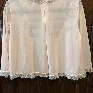 May include: A light pink, long-sleeved robe with a delicate lace trim along the collar, cuffs, and hem. The robe is hanging on a black hanger against a dark wood background. The robe has a simple, elegant design.