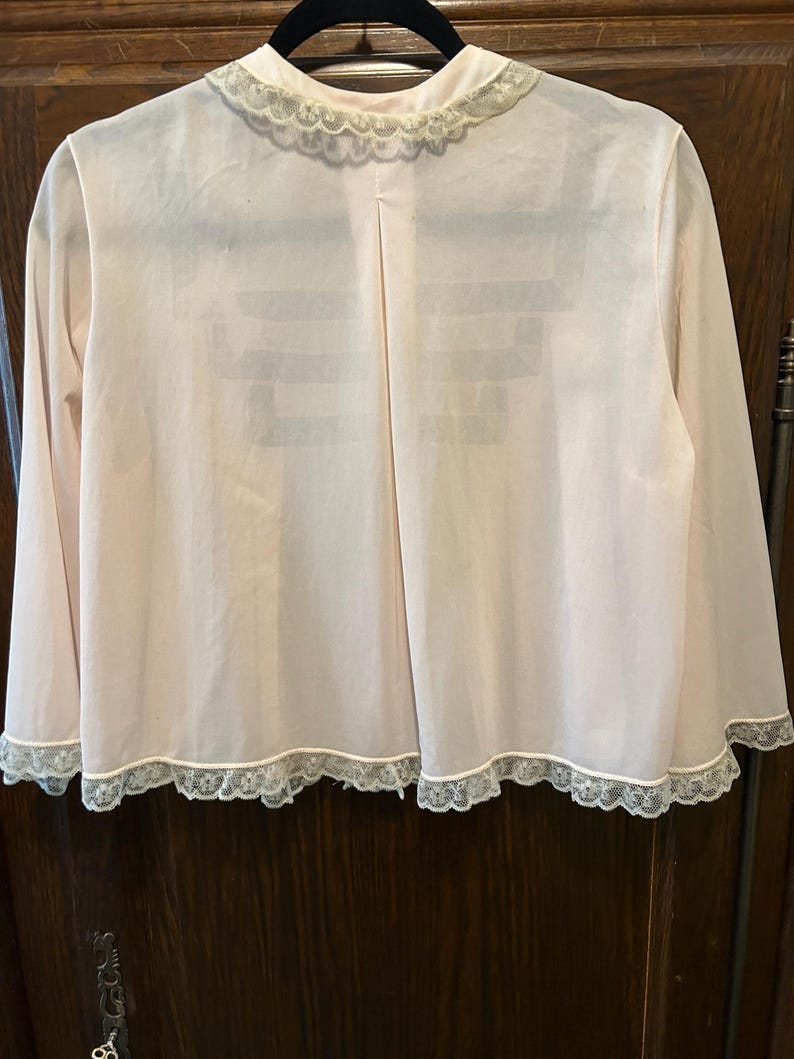 May include: A light pink, long-sleeved robe with a delicate lace trim along the collar, cuffs, and hem. The robe is hanging on a black hanger against a dark wood background. The robe has a simple, elegant design.