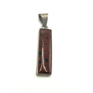 May include: A rectangular pendant featuring a smooth, polished reddish-brown stone with dark brown and black circular inclusions. The stone is set within a simple silver-toned metal frame and includes a triangular bail at the top for hanging.