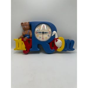 May include: A wall clock shaped like the words my room in blue, red, and yellow. A brown teddy bear sits on the letter m, and a white goose with a red bow sits on the letter o. The clock face is white with colorful numbers and black hands.