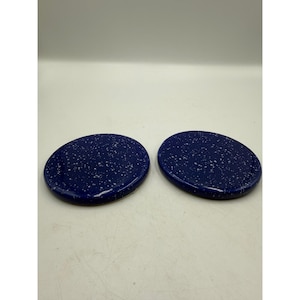 May include: Two oval-shaped, dark blue ceramic coasters with a speckled white pattern. The coasters are glossy and appear to be made of a durable material. They are designed to protect surfaces from heat and moisture.