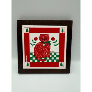 May include: Square decorative tile with a red cat design. The cat is covered in white polka dots and wears a green bow. The tile has a red and white border with green trees and flowers, set in a dark brown wooden frame.