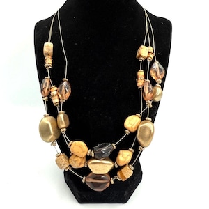 May include: A multi-strand necklace with a variety of beads in shades of brown, gold, and amber. The beads are of different shapes and sizes, including oval, round, and irregular. The necklace is displayed on a black mannequin.