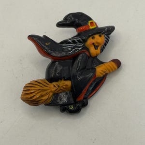 May include: A Halloween-themed brooch featuring a smiling witch riding a broom. The witch wears a black hat with a red band and a black cloak with red trim. The broom is brown, and the witch's face is orange.