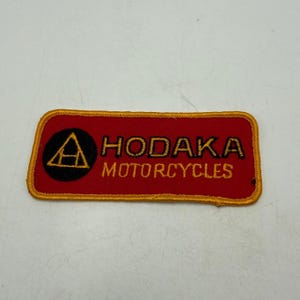 May include: A rectangular embroidered patch with a red background and a gold border. On the left is a black circle containing a gold triangle with an H inside. To the right, the text HODAKA MOTORCYCLES is embroidered in gold lettering.