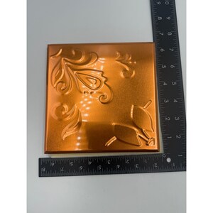 May include: A square, copper-colored decorative tile with embossed floral and scroll designs. The tile measures approximately 8 inches by 8 inches. The design features stylized leaves and swirling patterns, creating a textured surface.