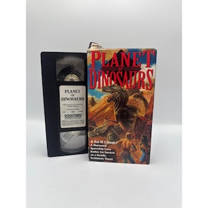 May include: A black VHS tape and its cardboard sleeve for the film Planet of Dinosaurs. The cover features a large dinosaur, rocky terrain, and two figures, with the title in red text. The tape label includes the title and Goodtimes Home Video logo.