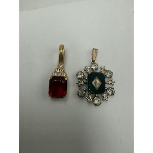 May include: Two gold-tone metal pendants. The left pendant features a rectangular red stone topped with small clear crystals. The right pendant is a circular starburst design with a central dark green rectangular stone and clear crystals.