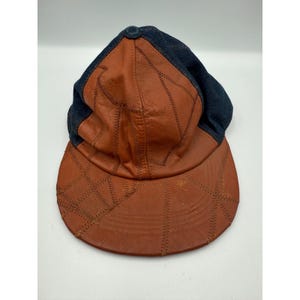 May include: A baseball cap with a brown leather visor and crown, and a dark blue denim top. The brown leather is stitched together in a patchwork design. A small, dark blue button is on top.