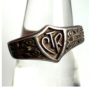 May include: A silver ring with a shield-shaped face featuring the letters "CTR". The ring has an ornate design with scrollwork and a textured band. The ring is likely made of sterling silver.