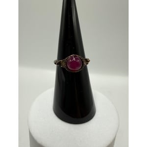 May include: A ring featuring a smooth, round, deep magenta cabochon stone set in a dark, textured metal band. The setting has a slightly weathered appearance with intricate detailing around the circular stone mount, creating a distinct contrast.