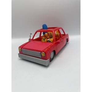 May include: A bright pink toy car featuring a silver front bumper and grille, with yellow headlights. Inside the vehicle, small yellow figures representing characters with blue hair and distinct facial features are seated in the front and back.