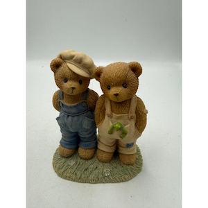 May include: A figurine of two brown teddy bears standing on a textured green base. The left bear wears a beige cap and blue overalls. The right bear wears cream-colored overalls with a small green frog tucked into the front pocket.