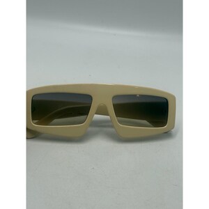 May include: Cream-colored sunglasses featuring a thick, angular frame with a flat top edge and rectangular lenses that have a dark gradient tint. The frame has a smooth, solid finish with visible hinges on the sides of the lens openings.