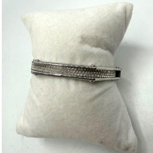 May include: A silver-toned hinged bangle bracelet featuring a central rectangular bar inlay encrusted with numerous small, clear, sparkling crystals. The bracelet has a polished metallic finish and a secure clasp mechanism on the side.