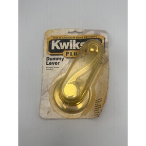 May include: A gold-colored Kwikset Plus dummy lever in its original packaging. The packaging features the text The #1 Choice in Home Security, Kwikset Plus, and Dummy Lever. The lever has a curved, ergonomic design and is mounted on a card.