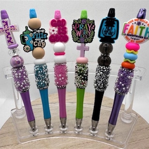 May include: A collection of decorative pens with various toppers, including crosses, inspirational quotes, and colorful beads. The pens feature purple, blue, pink, green, and black barrels, embellished with pearls and rhinestones. The pens are displayed in a clear acrylic stand.