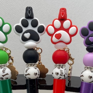 May include: Four colorful pens with paw print toppers. The pens have green, black, red, and purple paw prints with matching colored beads. Each pen has a charm and a bead with the words "Woof" and "Ruff" on it.