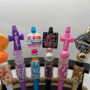 May include: A collection of decorative pens with inspirational messages. Each pen features a unique design, including crosses, circles, and text like "Blessed" and "She is Worthy." The pens have various colors, including purple, pink, and gold, and are embellished with beads and rhinestones.