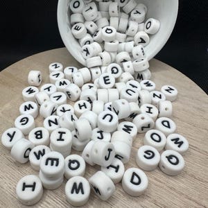 May include: White, round alphabet beads with black letters are scattered on a wooden surface, some spilling from a white bowl. The beads are cylindrical with a hole through the center, perfect for jewelry making or crafts.