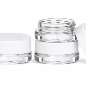 May include: Two small, clear glass cosmetic jars with smooth white screw-on lids. One jar is closed and sits to the left, while the other jar is open with its lid leaning against the side, revealing the transparent glass construction and thick base.