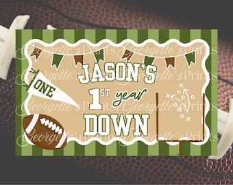 May include: A football-themed banner with the text "JASON'S 1st year DOWN." The design includes a football, a pennant that says "ONE," and a football field graphic. The background is a football.