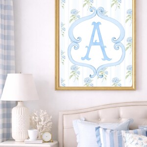 May include: A framed watercolor print featuring a light blue letter "A" within a decorative frame. The background has a floral and striped pattern in blue and white. The gold frame hangs above a bed with blue and white pillows.