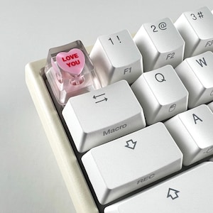 May include: Close-up of a mechanical keyboard with white keycaps. A custom keycap in the shape of a pink heart with the words "LOVE YOU" is featured. Other keys include "1!", "2@", "Macro", and "REC".
