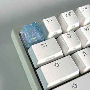 Puede incluir: Primer plano de un teclado de ordenador con una tecla personalizada. La tecla es azul translúcido con purpurina. Otras teclas son blancas con letras negras, incluyendo "1!", "2@", "Q" y "Macro".