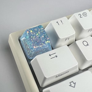 May include: Close-up of a keyboard with a custom keycap. The keycap is a translucent blue with embedded glitter. Surrounding keys are white with black lettering, including "1!", "2@", "F1", and "Macro".