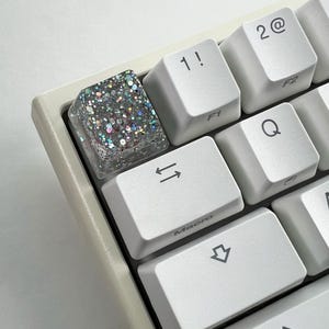 Pode incluir: Close-up de um teclado de computador com uma tecla personalizada. A tecla é transparente com glitter prateado e iridescente embutido. Outras teclas são brancas com letras e símbolos pretos, incluindo "1!", "2@" e "Macro".