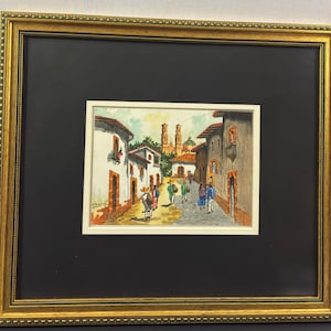 May include: Watercolor painting of a street scene in a Mexican town, framed in gold. The artwork depicts buildings with red tile roofs, a church in the background, and people walking on the cobblestone street. The painting is signed in the lower left corner.