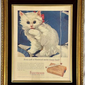 May include: Framed vintage advertisement featuring a fluffy white cat with a red bow, sitting on a blue and white checkered surface. The ad promotes Fleetwood cigarettes, with text that reads "Every puff of Fleetwood smoke Cleans Itself!"