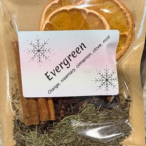 May include: A clear plastic bag filled with dried herbs, spices, and orange slices. The label reads "Evergreen" with the ingredients: orange, rosemary, cinnamon, clove, and mint. The bag is sealed and set against a brown paper background.