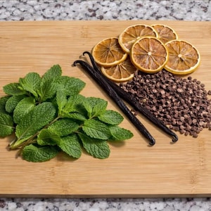 Può includere: Un tagliere in legno mostra menta fresca, fette di arancia essiccate, gocce di cioccolato fondente e due baccelli di vaniglia. Gli ingredienti sono disposti sulla tavola, suggerendo un tema culinario. La tavola è su un piano di lavoro maculato.