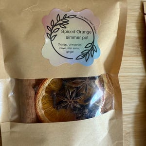 Könnte beinhalten: Ein brauner Papiersack mit einem transparenten Fenster, das einen Spiced Orange Simmertopf zeigt. Das Etikett lautet "Spiced Orange Simmertopf" mit den Zutaten: Orange, Zimt, Nelken, Sternanis und Ingwer. Der Beutel enthält getrocknete Orangenscheiben, Zimtstangen und Sternanis.