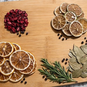 Könnte beinhalten: Ein hölzernes Schneidebrett mit verschiedenen getrockneten Zutaten. Es gibt Haufen von getrockneten Cranberries, Limettenscheiben, Zitronenscheiben, Lorbeerblättern, Rosmarinzweigen und Pfefferkörnern. Die Zutaten sind in separaten Abschnitten angeordnet.