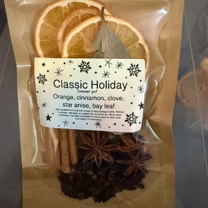 May include: A clear bag containing dried orange slices, cinnamon sticks, star anise, cloves, and a bay leaf. The label reads "Classic Holiday" and lists the ingredients. The bag is sealed. The ingredients are for a simmer pot.