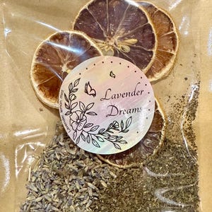 May include: A clear plastic bag containing dried lemon slices, lavender buds, and dried mint. A round sticker with the words "Lavender Dreams" and floral illustrations is centered on the bag. The bag is sealed and set against a brown paper background.