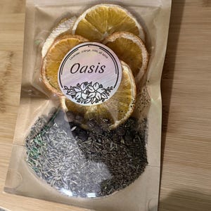 Op de afbeelding: Een transparante zak gevuld met gedroogde sinaasappelschijfjes, kruiden en specerijen. Een rond etiket met het woord "Oasis" en de tekst "Lavendel, sinaasappel, munt, alle kruiden" is zichtbaar. De zak is verzegeld en ligt op een houten oppervlak.