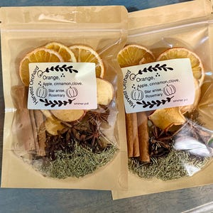 Może przedstawiać: Dwa przezroczyste woreczki wypełnione suszonymi plasterkami pomarańczy, kawałkami jabłek, laskami cynamonu, anyżem gwiazdkowatym, goździkami i rozmarynem. Każdy woreczek ma etykietę z napisem "Harvest Orchard" i zawiera listę składników: pomarańcza, jabłko, cynamon, goździk, anyż gwiazdkowaty i rozmaryn.