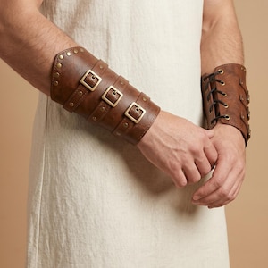 May include: A pair of brown faux leather bracers featuring three horizontal straps with brass-toned metal buckles and studs. The bracers are secured with black lacing through metal eyelets along the side, providing an adjustable fit.