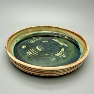 May include: A handmade ceramic serving dish with a circular shape. The dish features a dark teal interior with concentric circle patterns and a light tan rim with brown stripes. The overall aesthetic is rustic and artistic.