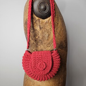 Könnte beinhalten: Eine handgefertigte, rote Häkeltasche mit Klappenverschluss und langem Riemen. Die Tasche hat einen gewellten Rand und ein kreisförmiges Knopfdetail auf der Klappe. Der Riemen ist ebenfalls gehäkelt und mit silberfarbenen Ringen an der Tasche befestigt.