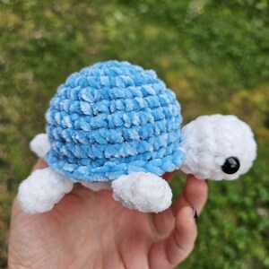May include: A handmade, plush turtle toy with a light blue shell and white head, feet, and tail. The turtle has a black eye and is made of soft, textured yarn. The toy is designed for children.