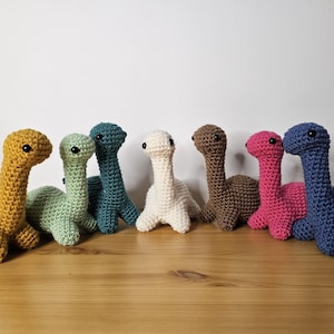 Könnte beinhalten: Eine Sammlung von sieben gehäkelten Dinosaurierspielzeugen in verschiedenen Farben, darunter Gelb, Grün, Türkis, Weiß, Braun, Rosa und Blau. Jeder Dinosaurier hat einen langen Hals und kleine schwarze Augen, auf einer Holzoberfläche angeordnet.