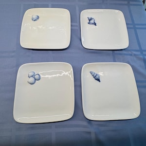 Puede incluir: Cuatro platos cuadrados blancos con esquinas redondeadas, cada uno adornado con un diseño de concha azul. Los platos están dispuestos sobre un mantel azul, lo que sugiere un tema costero. Los diseños de conchas varían ligeramente.