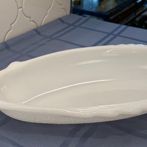 Könnte beinhalten: Eine weiße, ovale Servierschale mit strukturierten Kanten und Griffen. Die Schale besteht aus glänzendem Material und steht auf einer blauen Tischdecke. Die Schale ist leer und bereit, mit Essen gefüllt zu werden.